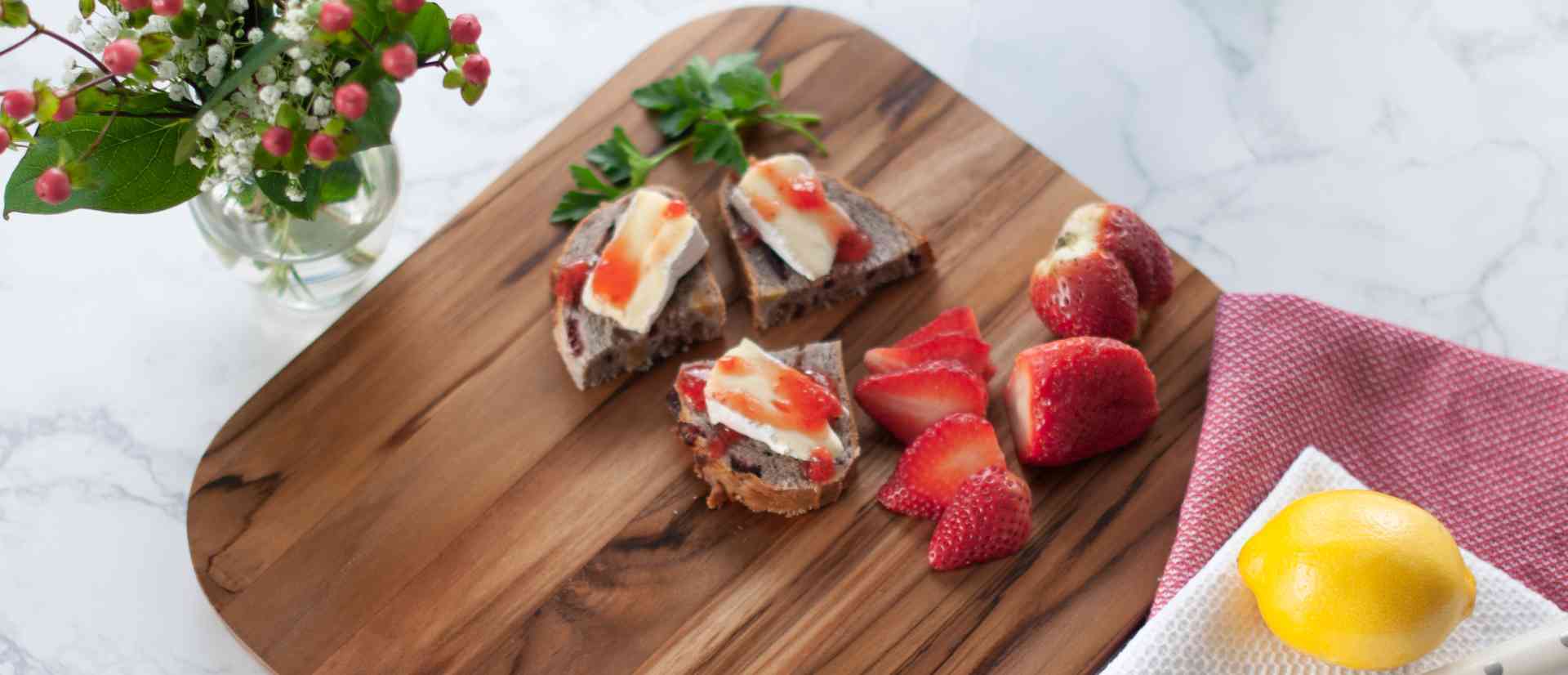 A teak cutting board with strawberries and bread with brie
